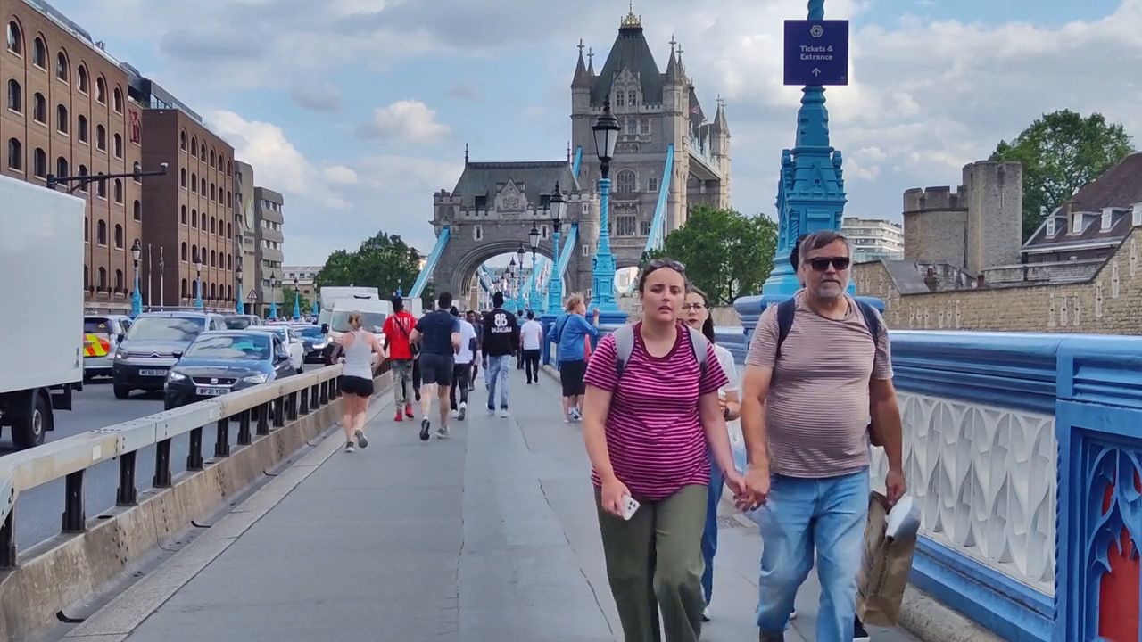 People walking along the footway of Tower Bridge, with blue railings and a stone tower ahead