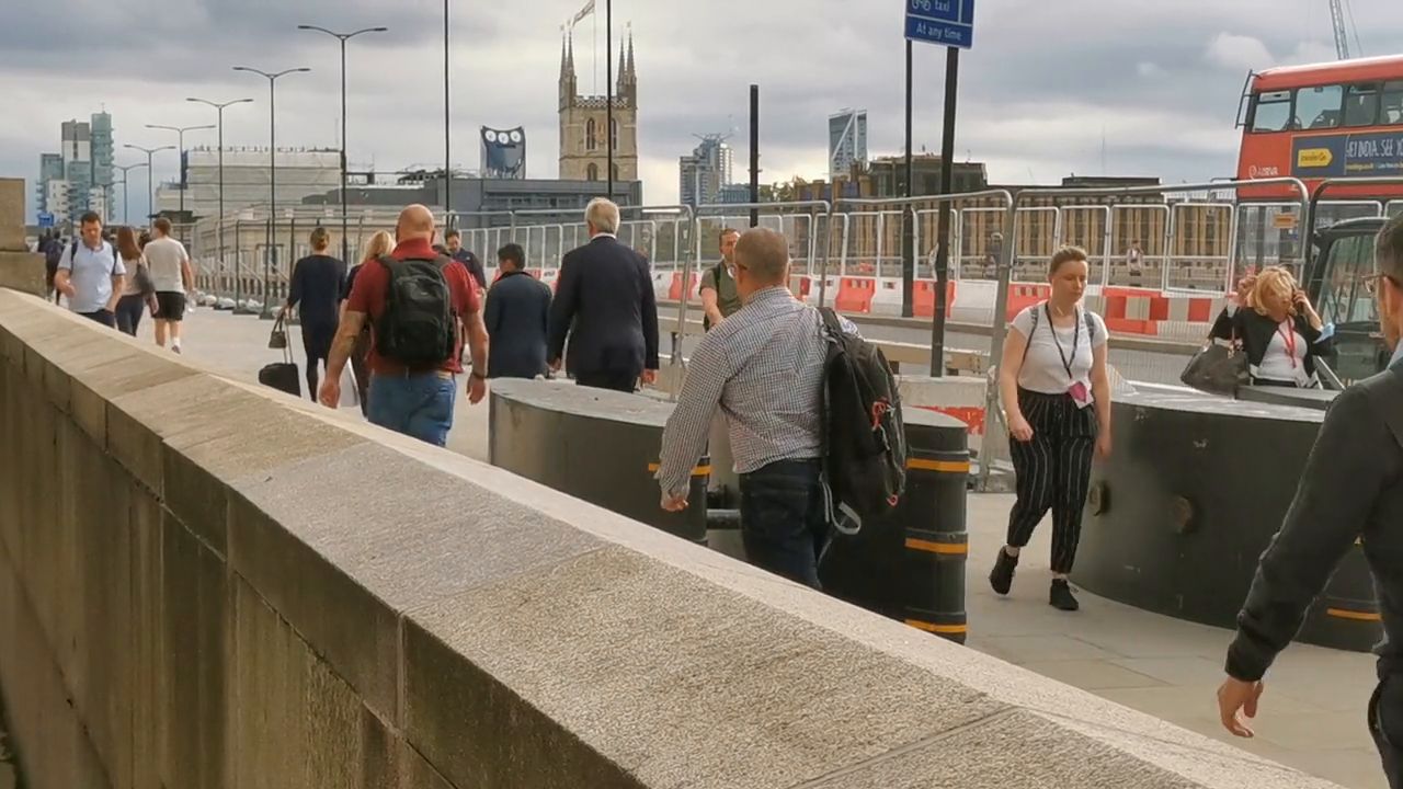 Pedestrians walking beside the stone parapet of London Bridge, with traffic barriers on the left