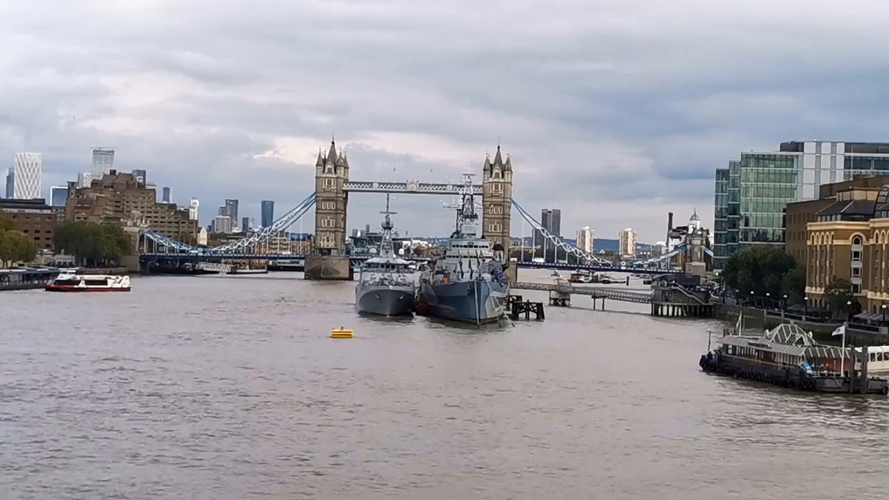 A wide grey stretch of the Thames with Tower Bridge small in the distance and boats moored along the bank