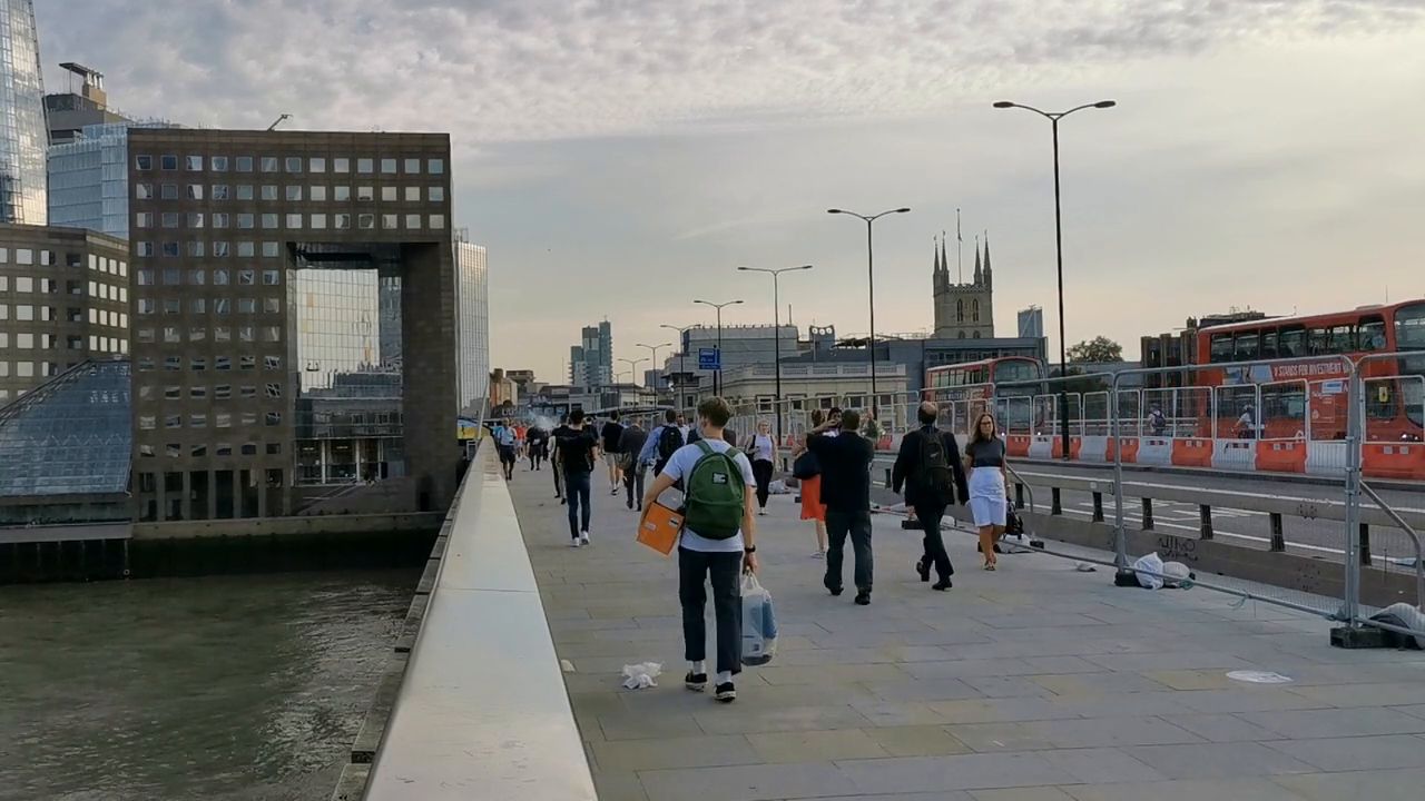 People walking on London Bridge, with lamp posts, a low barrier and the City skyline behind